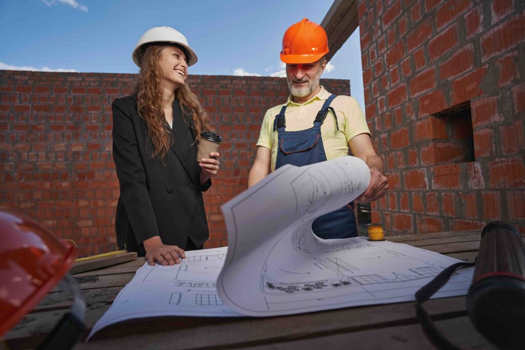 Female engineer with coffee looking at construction worker with smile while he inspecting house layout on table