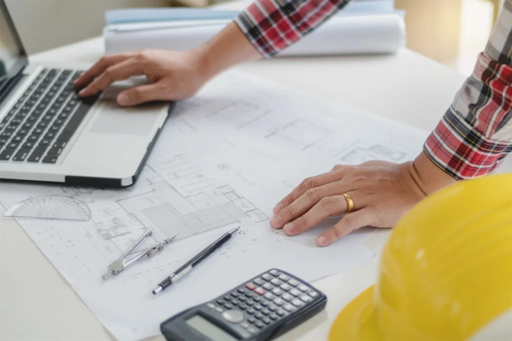 MEP drafting virtual assistant working on construction plans beside laptop, calculator, and hard hat